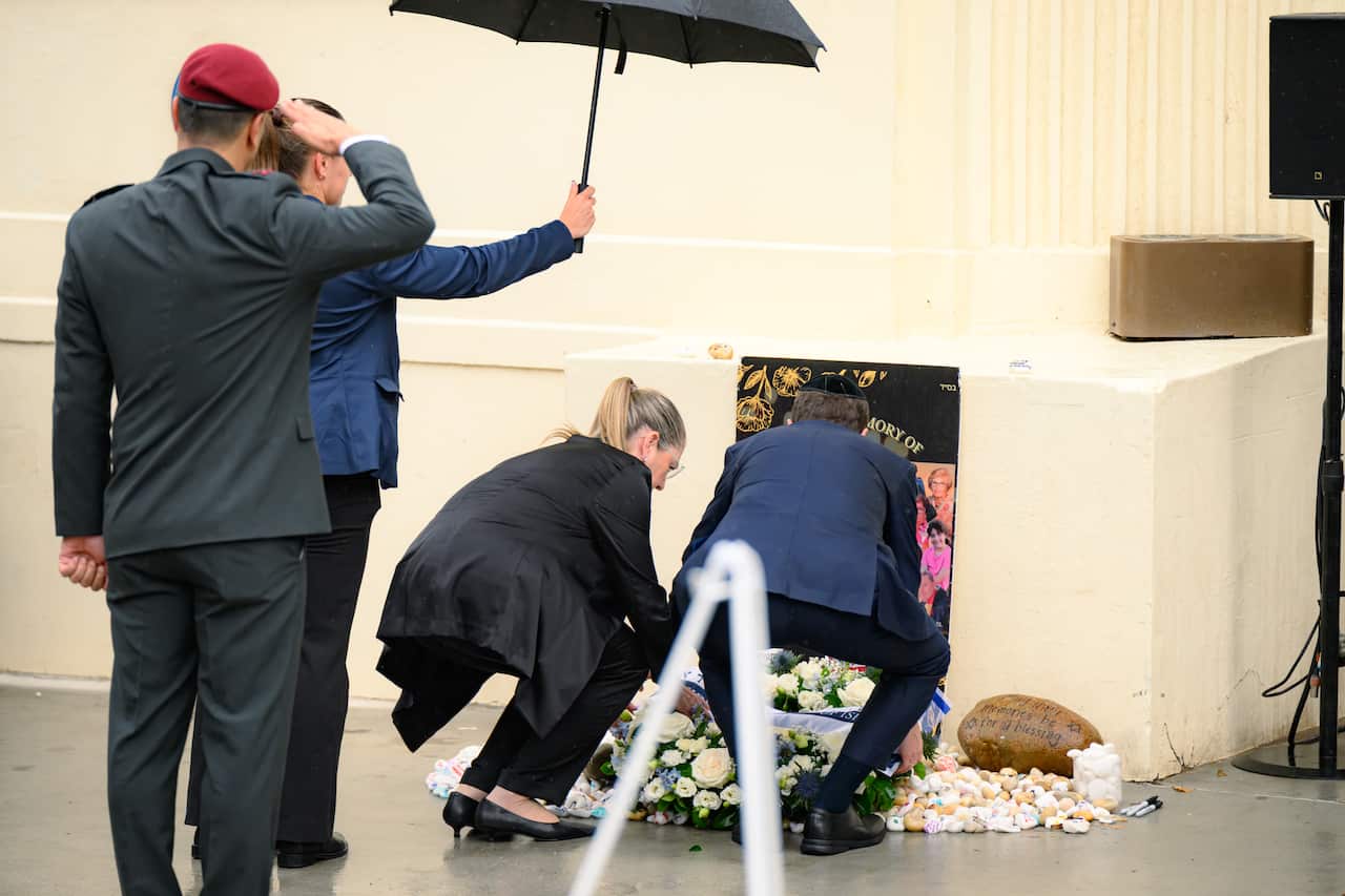 A man and woman laying a wreath of flowers. A woman holds an umbrella over them while a man with a red beret salutes them. 