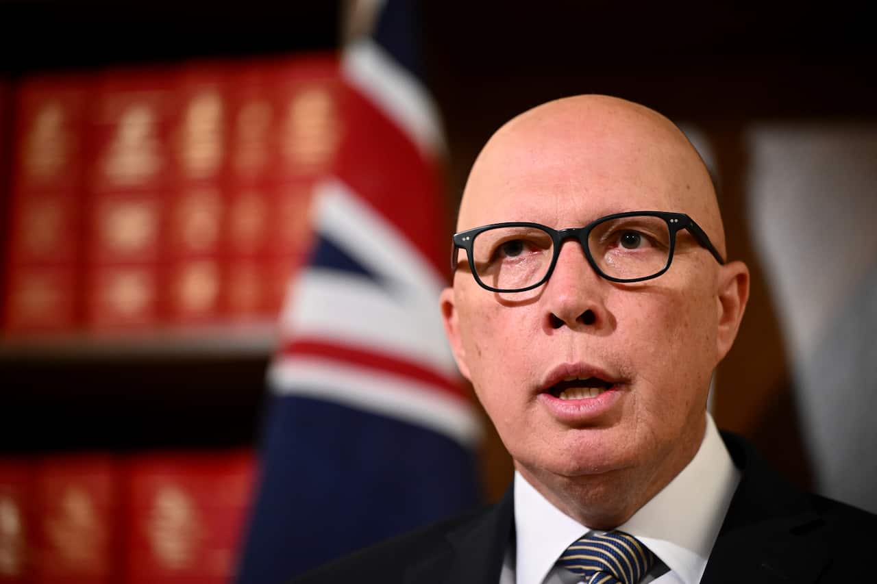 A bald man wearing glasses with his mouth open with an Australian flag and books in the back drop