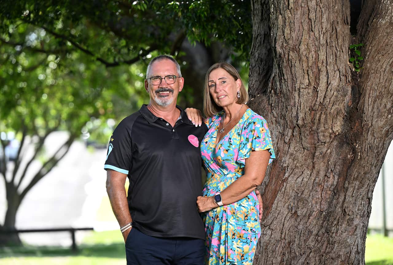 A man and a woman in a half embrace are standing outside in front of a tree.
