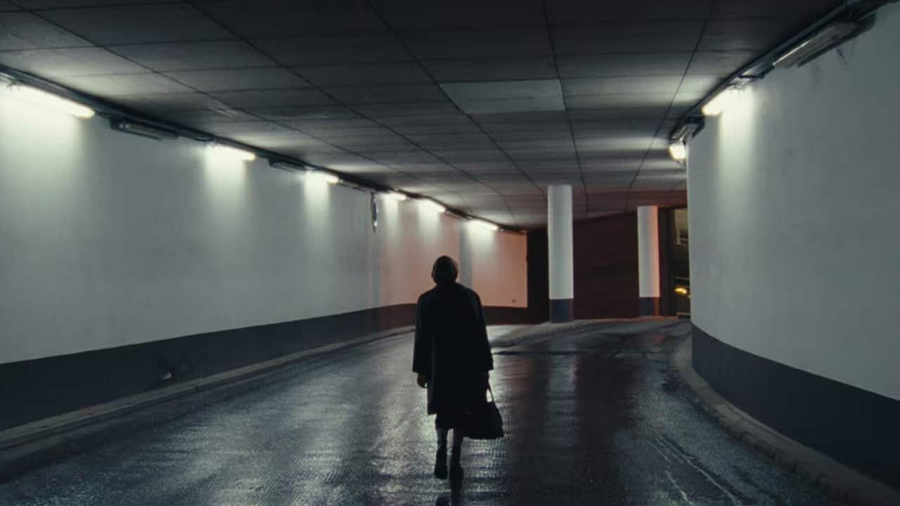 A figure in dark clothing walks along a curving path in what looks to be an underground location. Although the walls are white, the wide path and ceiling are dark, and the lights are dim, creating a lonely mood. 