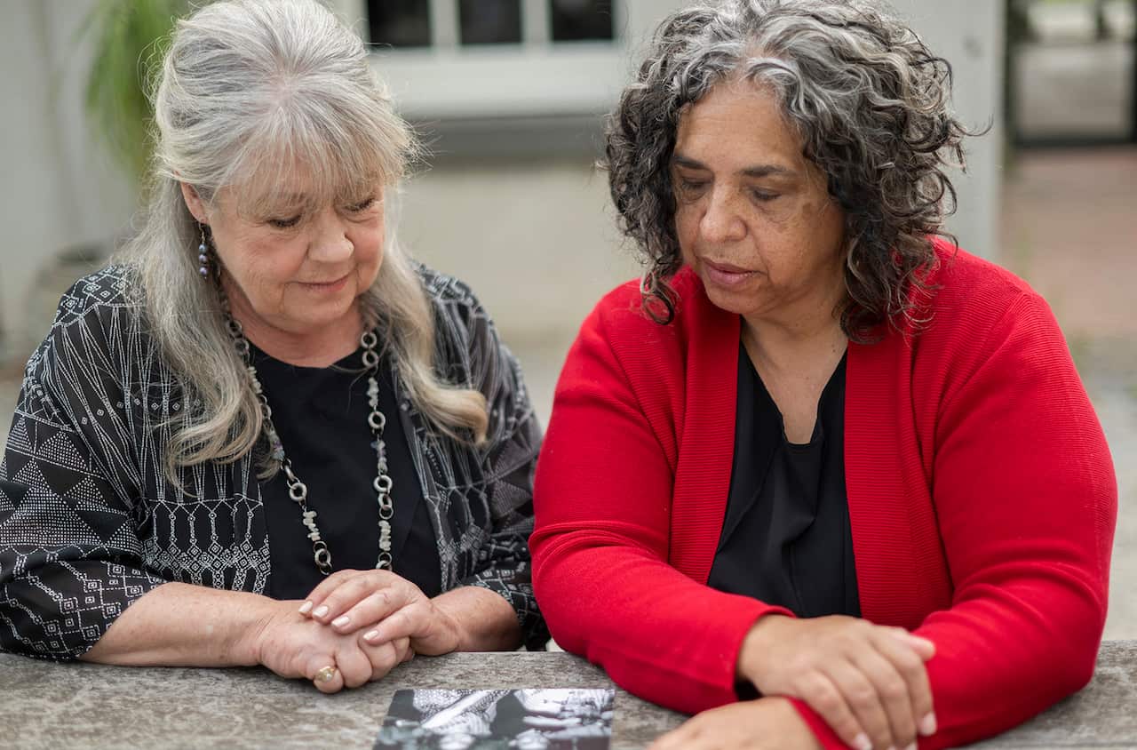 Two women sit at a marble bench, looking down at a black and white photograph.