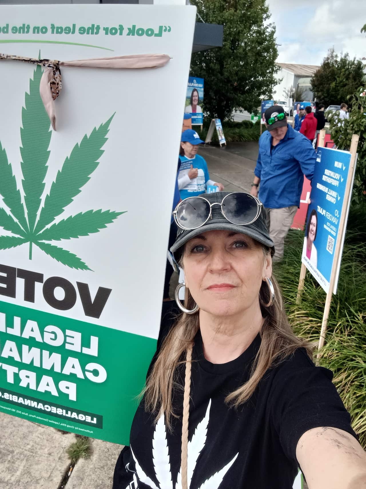A woman wearing a black shirt with a white marijuana leaf holds up a Legalise Cannabis Party corflute at a polling station.