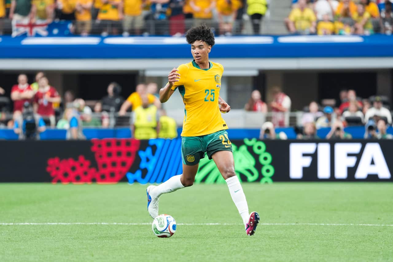 A footballer wearing a yellow shirt and green shorts controls the ball on the pitch during a match with fans in the background.