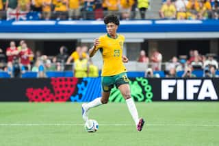 A footballer wearing a yellow shirt and green shorts controls the ball on the pitch during a match with fans in the background.
