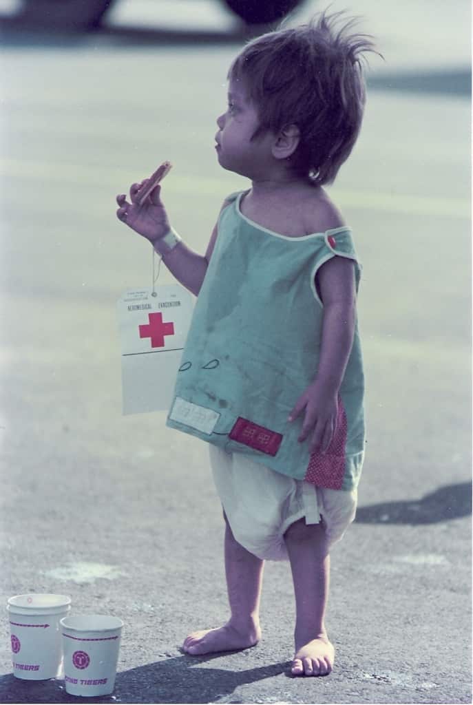 A three-year-old girl standing on an airport tarmac wearing a nappy, green singlet, red cross tag on her wrist, eating a biscuit.
