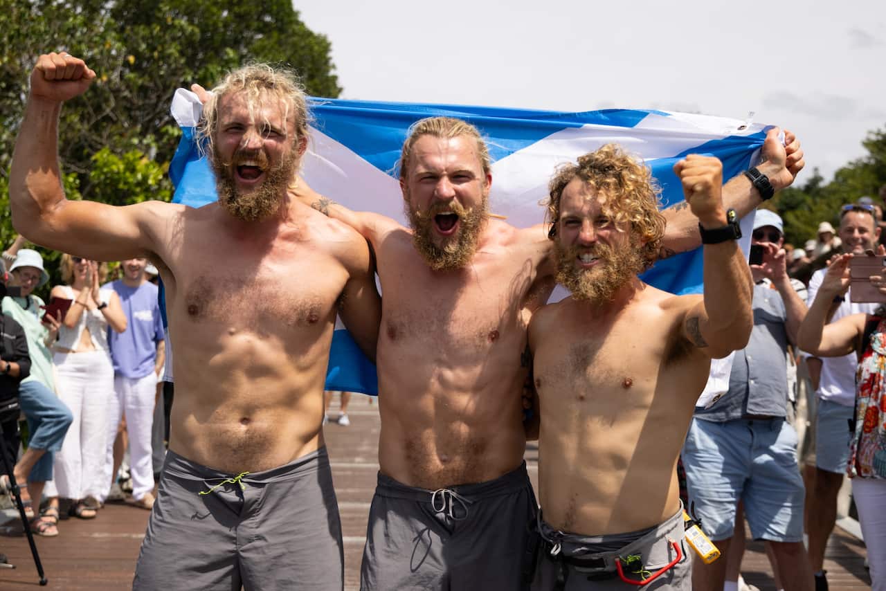 Three men with their fists raised and cheering, standing in front of a Scottish flag.