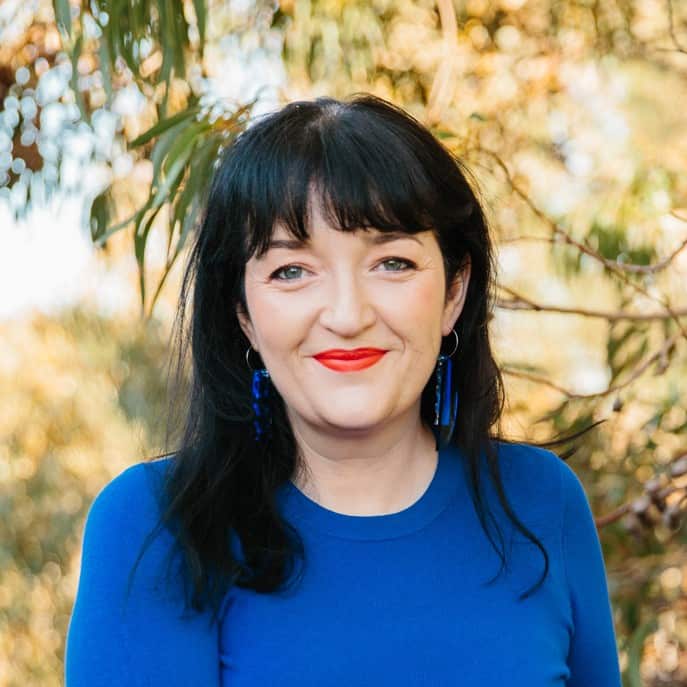 A woman with black hair, wearing a blue top and earrings and red lipstcik