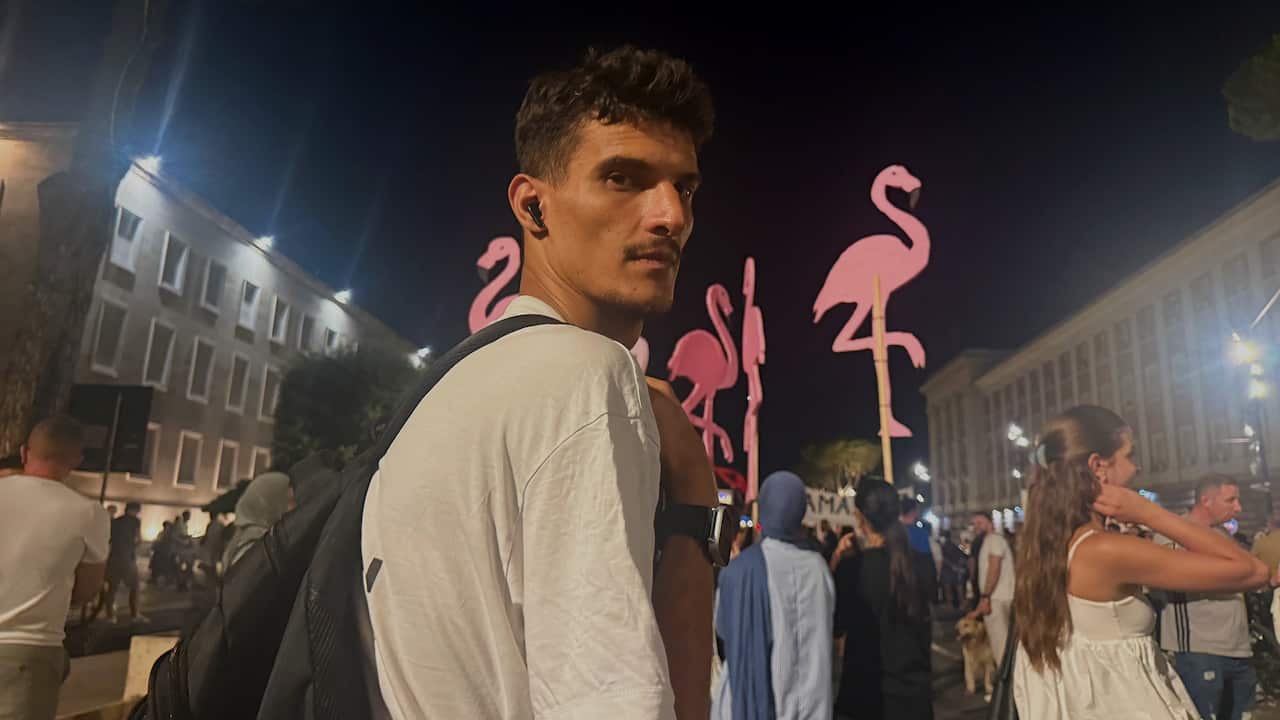 A young man wearing a white T-shirt and backpack looks over their shoulder while standing in a crowd at a nighttime gathering in a city square. Several large pink flamingo-shaped signs rise above the crowd