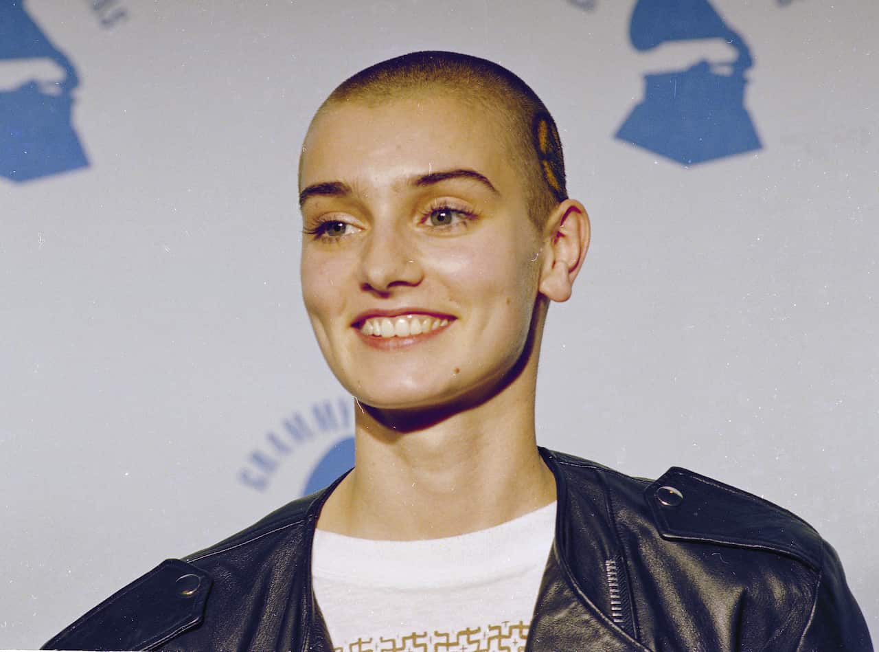 Woman with shaved head and black leather jacket smiles as she stands in front of an award show sign.