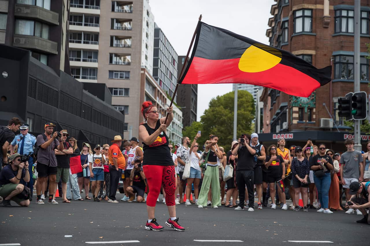 A woman wearing a black top and red leggings waves an Aboriginal flag at a rally.