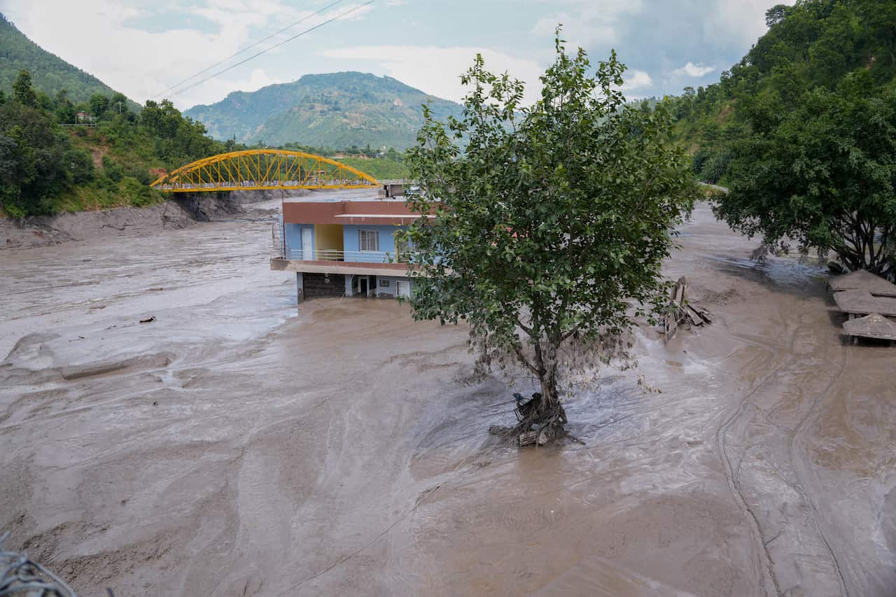 Mud and floodwater surround a partly submerged building and a tree in a river valley, with a yellow bridge, forested hills and extensive sediment deposits visible in the background.