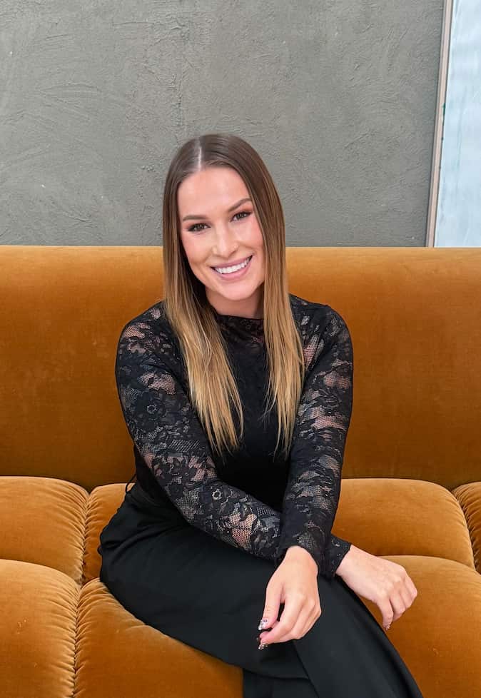 A smiling woman with long hair and a black, long sleeve lace top sits on a brown velvet couch.