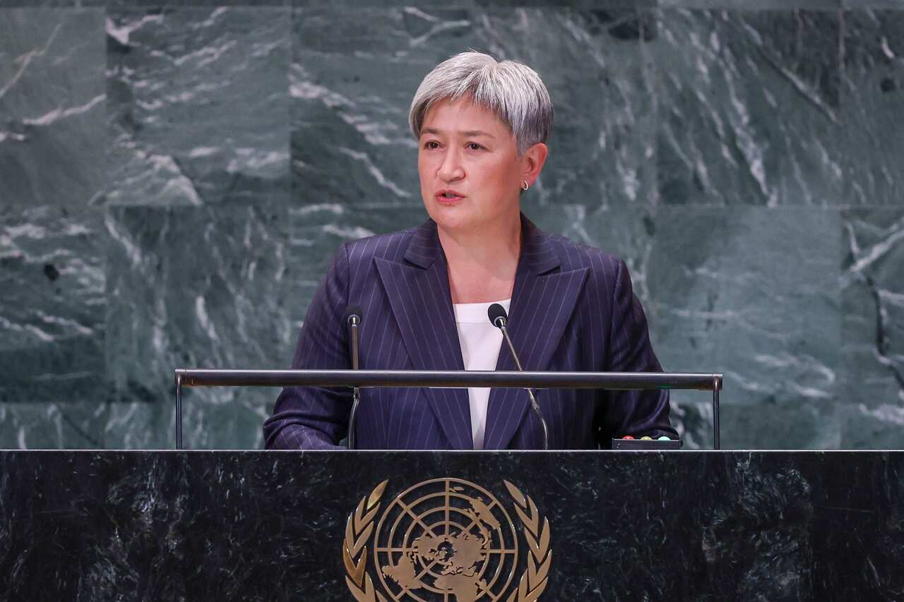 A woman in a dark striped suit and short silver hair stands at a marble podium. 