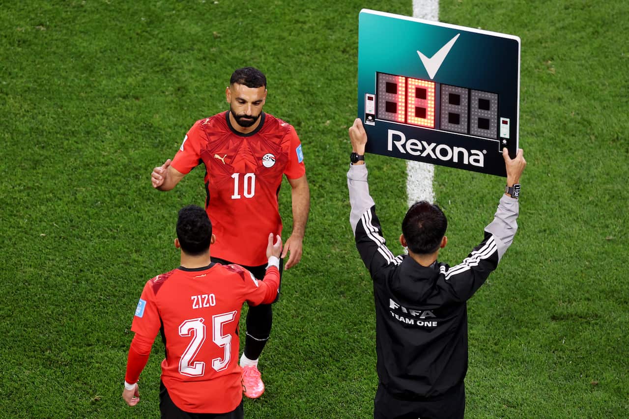 Egypt captain Mohamed Salah walks onto the pitch as a substitute during a FIFA World Cup match, reaching out to greet teammate Zizo as he comes off the field. A fourth official stands beside them holding an electronic substitution board above his head.