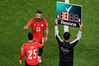 Egypt captain Mohamed Salah walks onto the pitch as a substitute during a FIFA World Cup match, reaching out to greet teammate Zizo as he comes off the field. A fourth official stands beside them holding an electronic substitution board above his head.