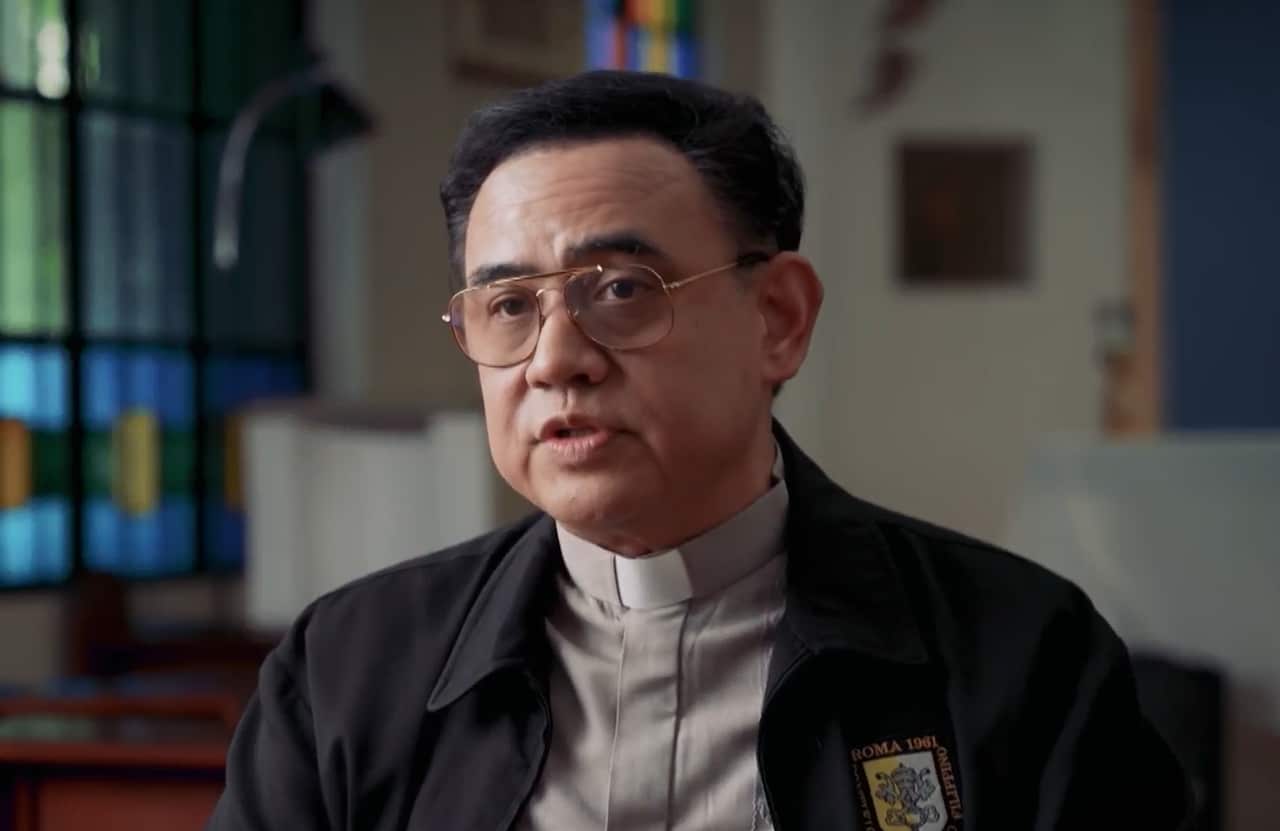 A priest with dark hair and glasses sits in a church. 