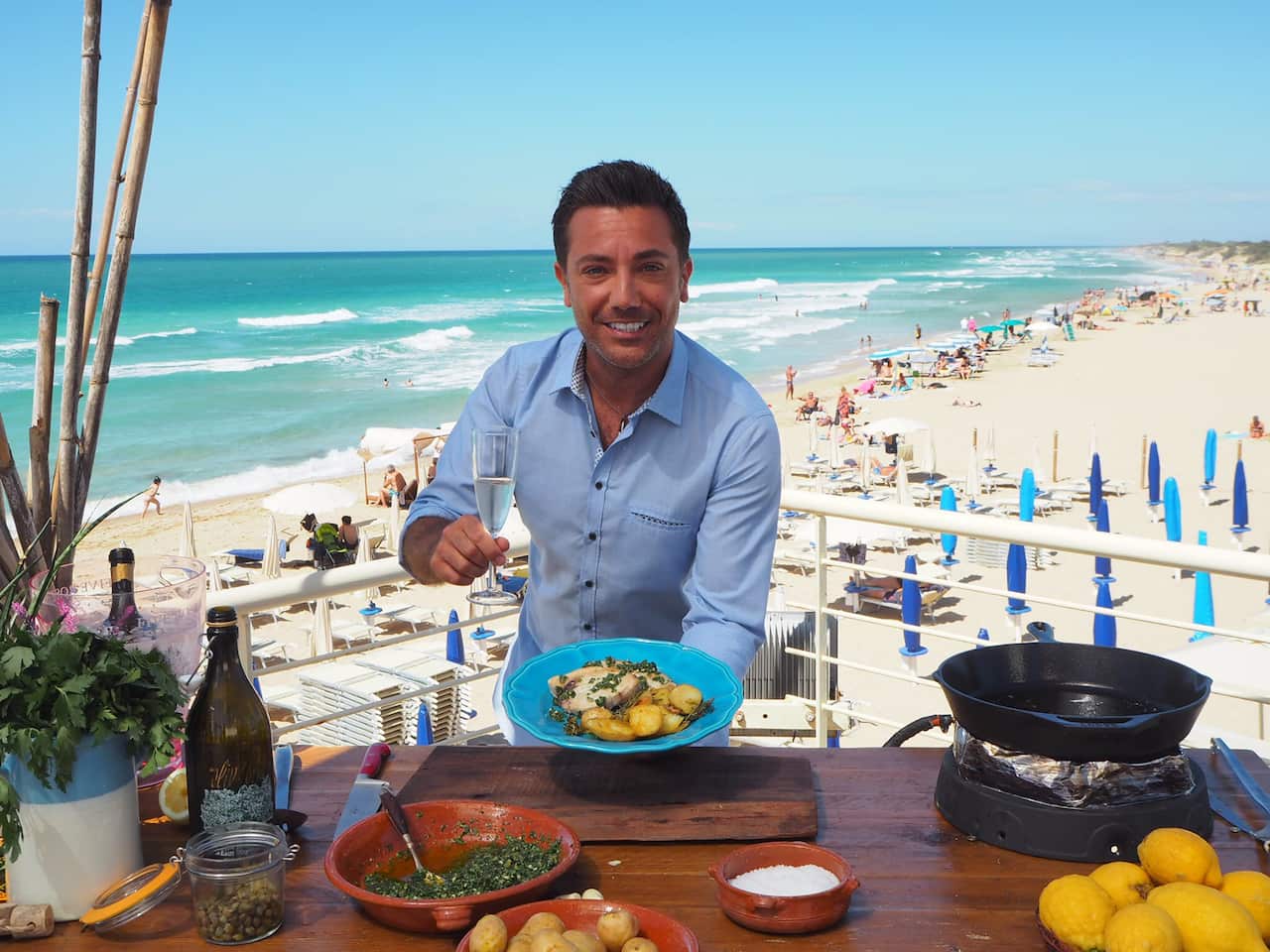 Gino D'Acampo stands behind a table, holding a glass of sparkling wine and a plate of food. A long sunny beach stretches away into the distance behind him.