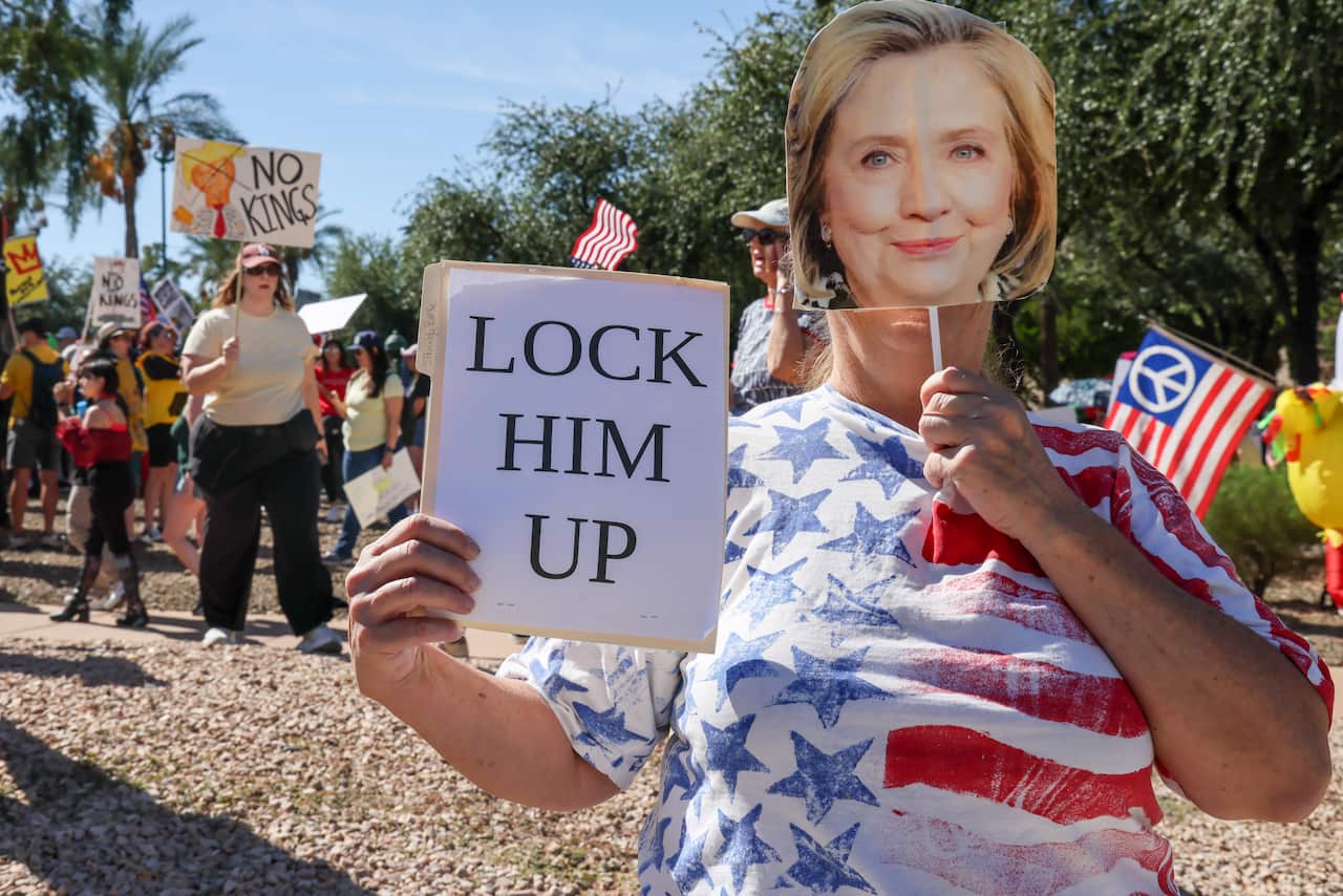 A protestor in a stars-and-stripes shirt holds a sign that reads "LOCK HIM UP" and a cutout mask of Hillary Clinton's face, while other protestors in the background hold signs that say "NO KINGS" and feature a cartoon of Donald Trump.