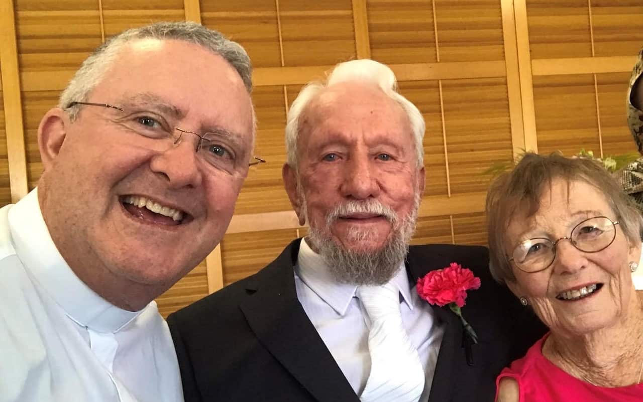 A man in a white shirt and clerical collar standing next to another, elderly man in a black suit and an elderly woman in a pink dress. All three are smiling.