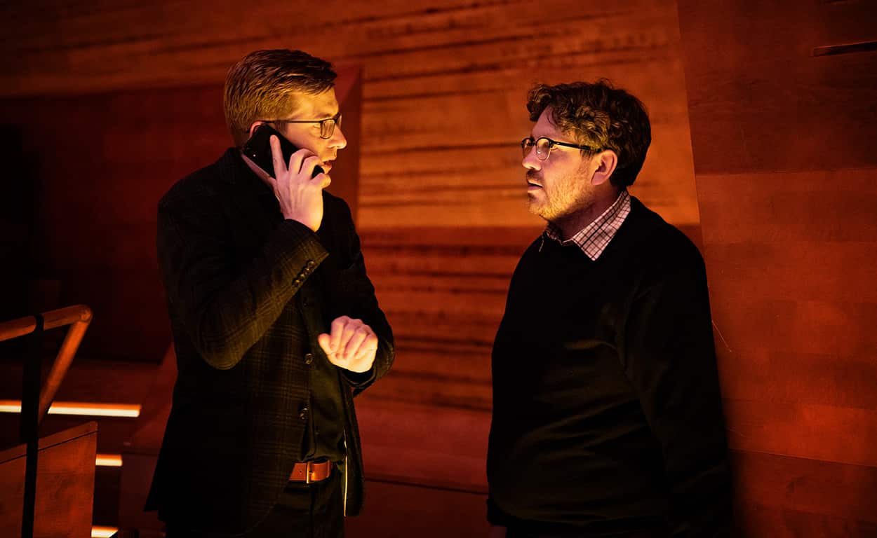 Two men stand beside a wood-panelled wall, in dim light (suggestive of a concert hall). One is making a phone call.