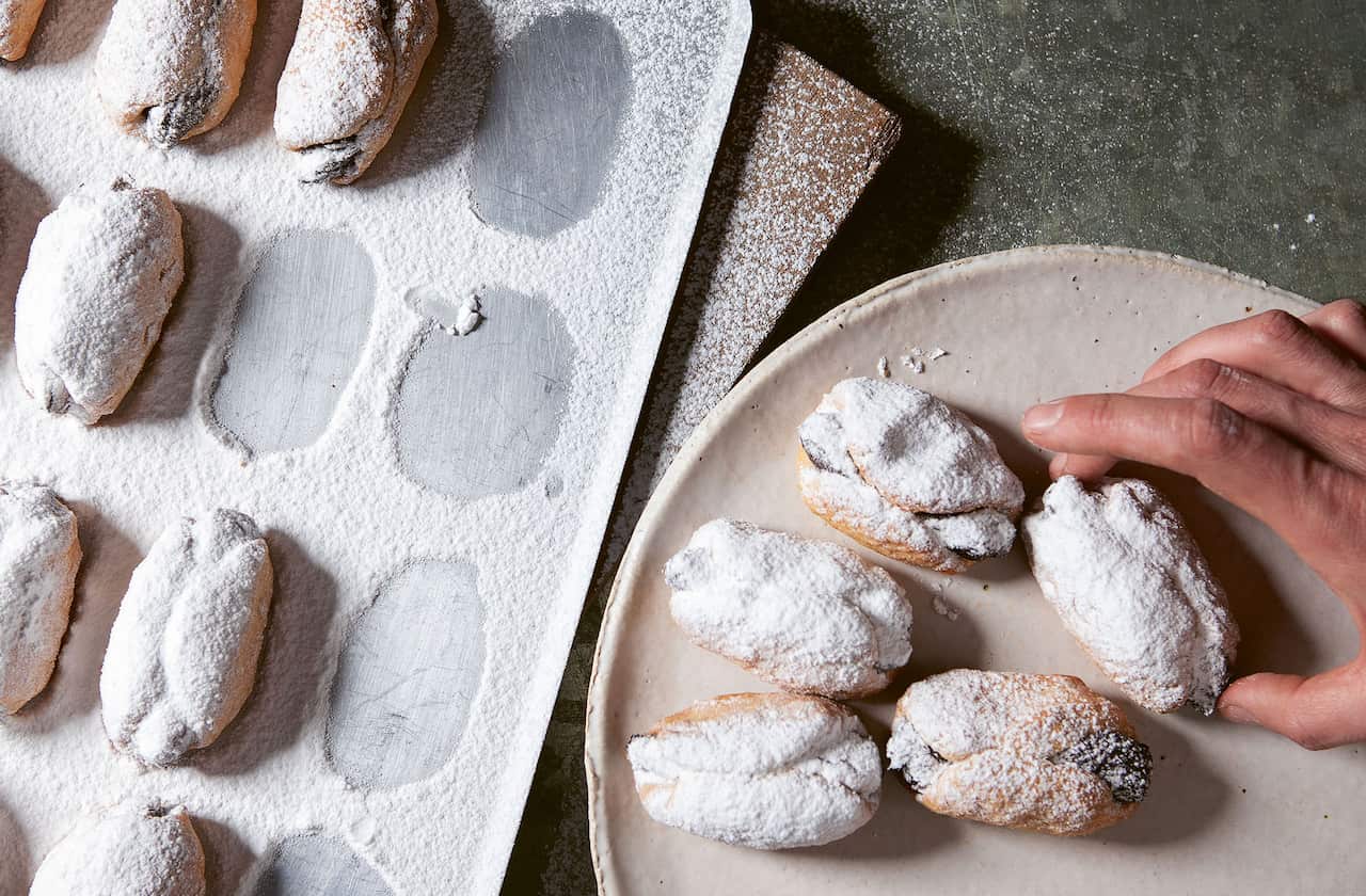 A tray of icing-sugar dusted oval biscuits sits beside a round plate. A hand is placing a biscuit, lifted from the tray, onto the plate. 