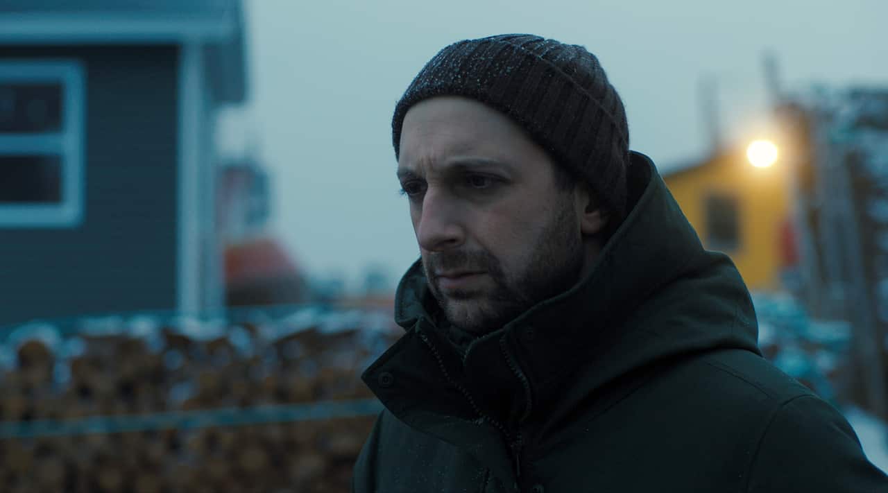 A man in a dark caot and beanie stands in a dim outdoor setting. Buildings can be seen, our of focus, behind him. 