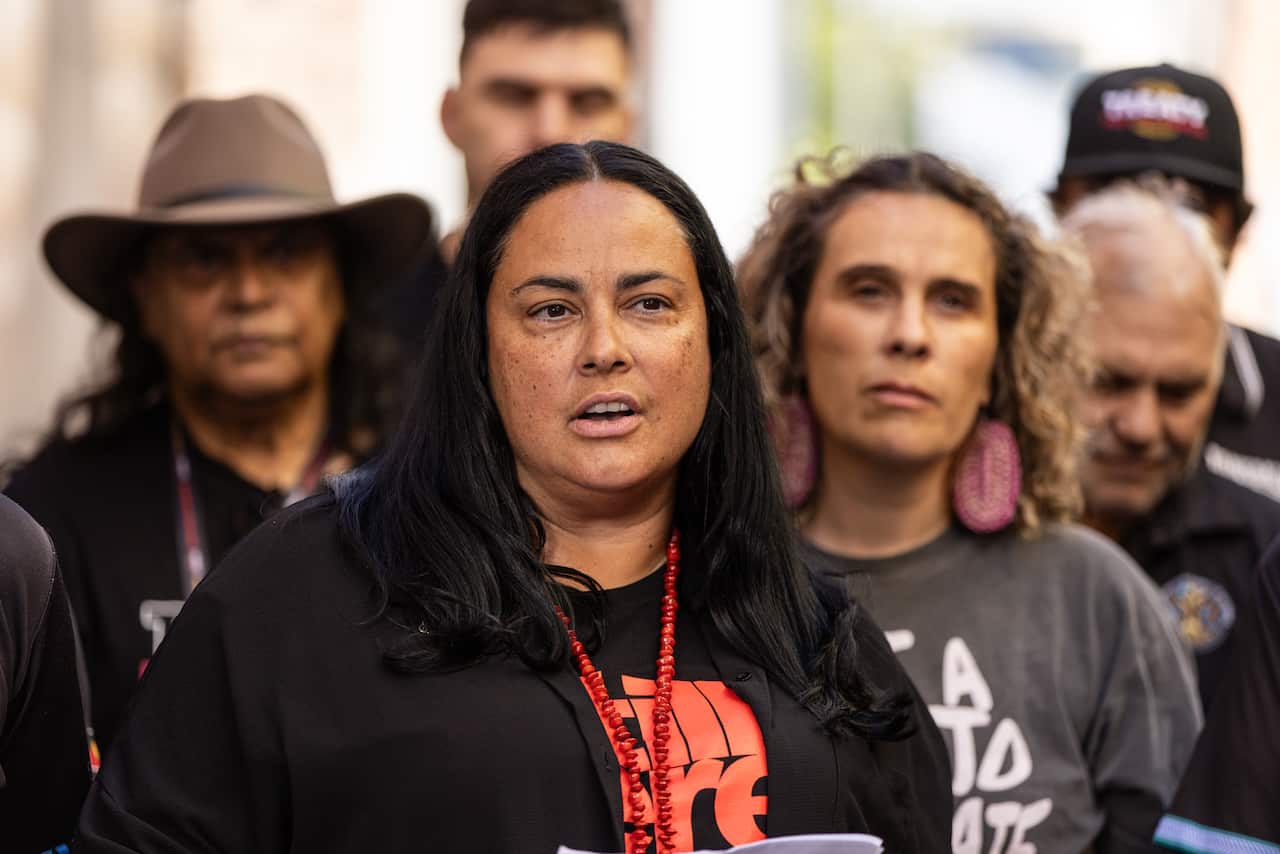 A woman with long black hair speaking, with others behind her.