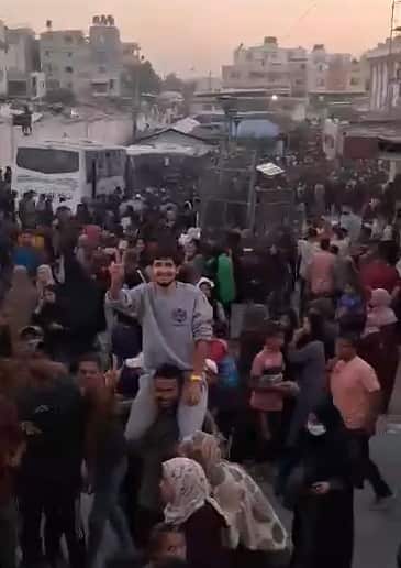 A dense crowd of people is gathered in a street amidst buildings and a bus, with a young man smiling and holding up a peace sign while being carried on another person's shoulders.