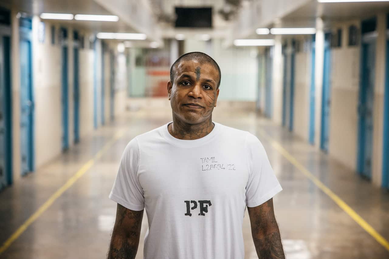 A prisoner in a white T-shirt with some facial tattoos look into the camera. Behind him are the blue doors of Changi prison cells.