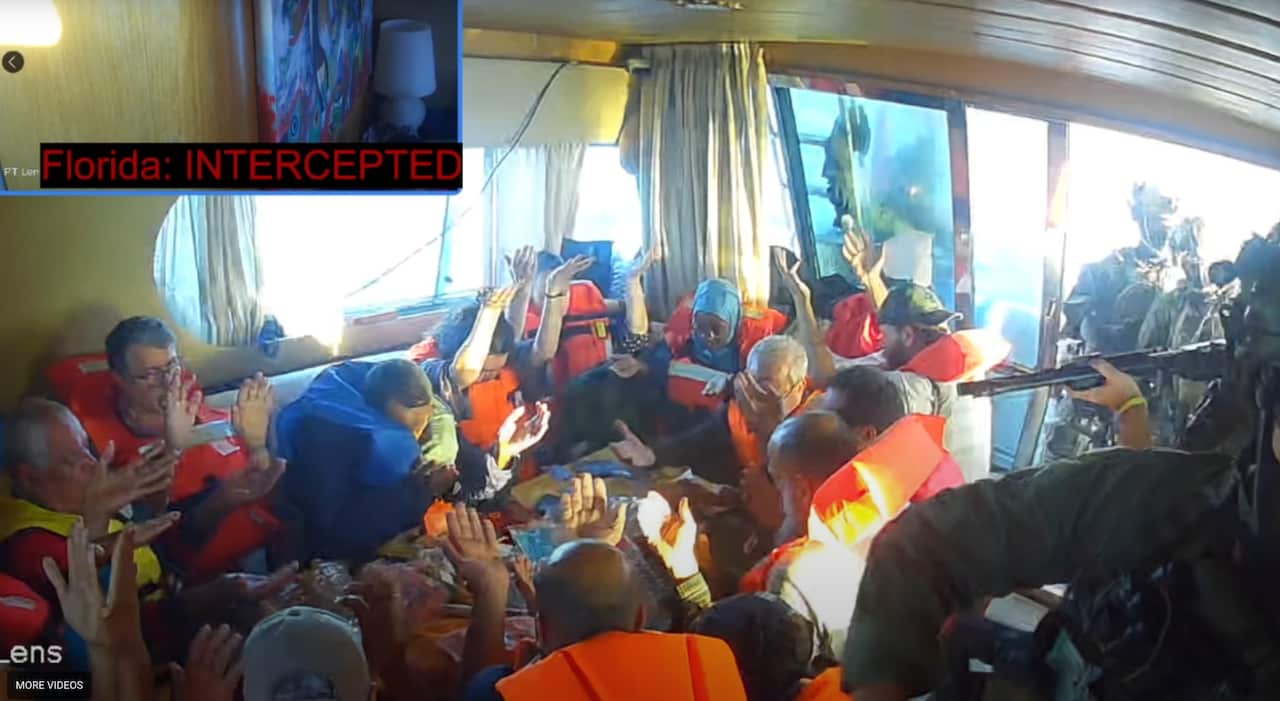 People in orange life jackets huddled in a circle with their hands in the air. THey are surrounded by figures in dark clothes holding guns.