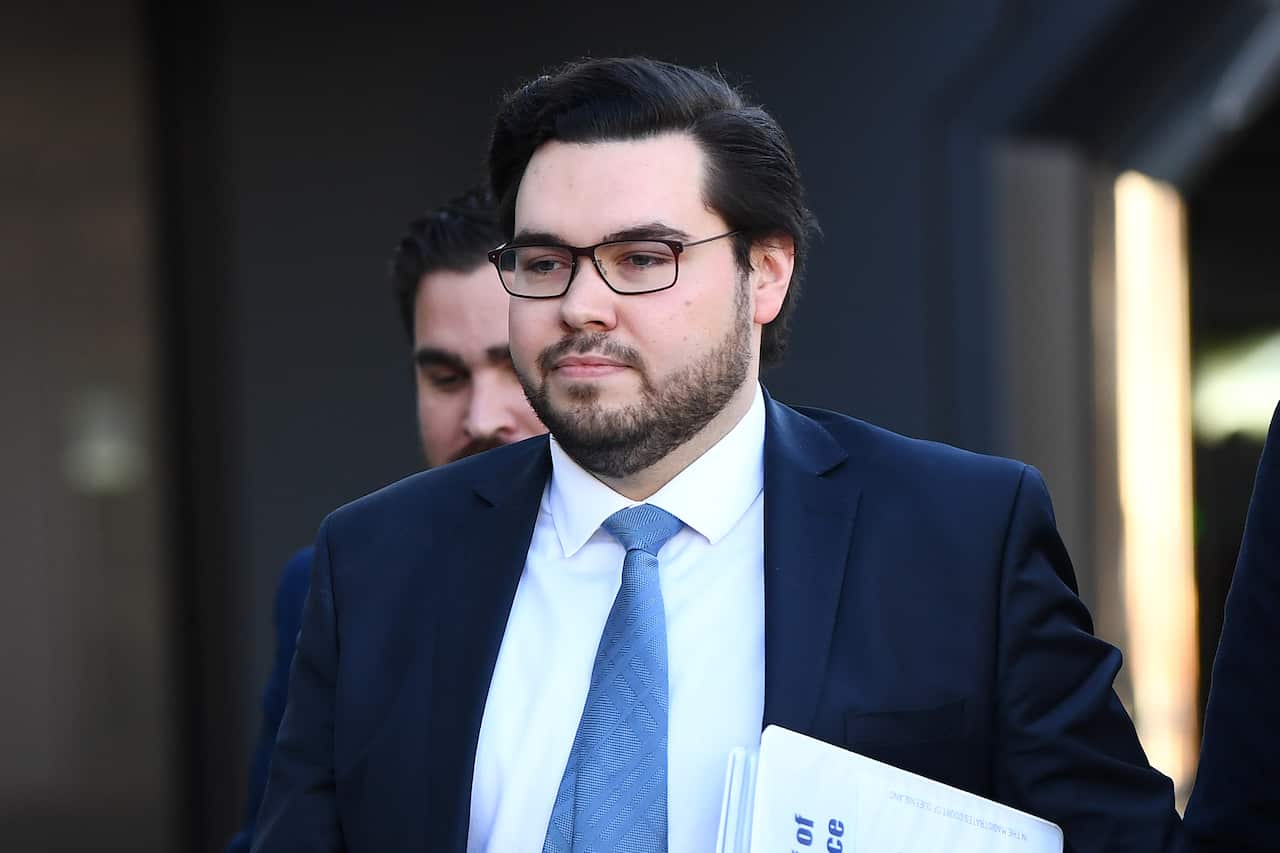 A man with dark hair and glasses wearing a dark blue suit and light blue tie walks forward 