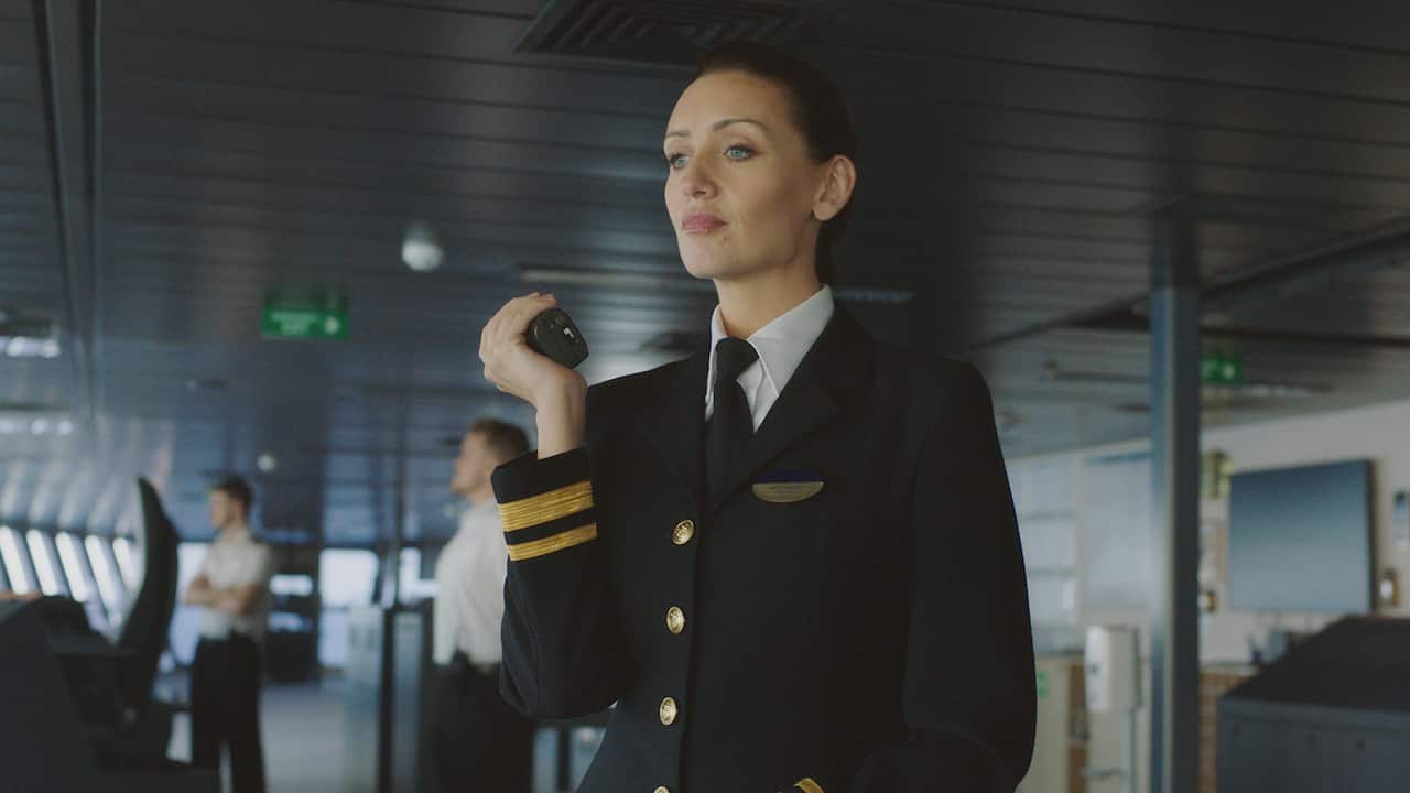 A woman in a formal ship's officer jacket, white shirt and tie stands on a deck, holding a radio.