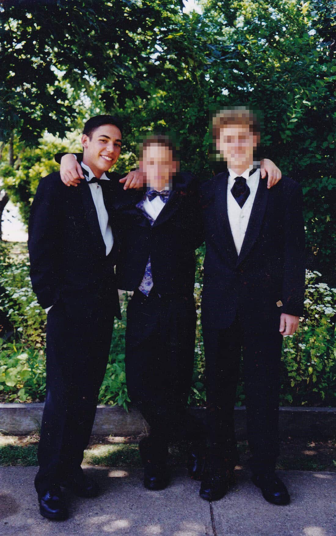 Three young men in formal black suits standing outside. They have their arms over each other's shoulders