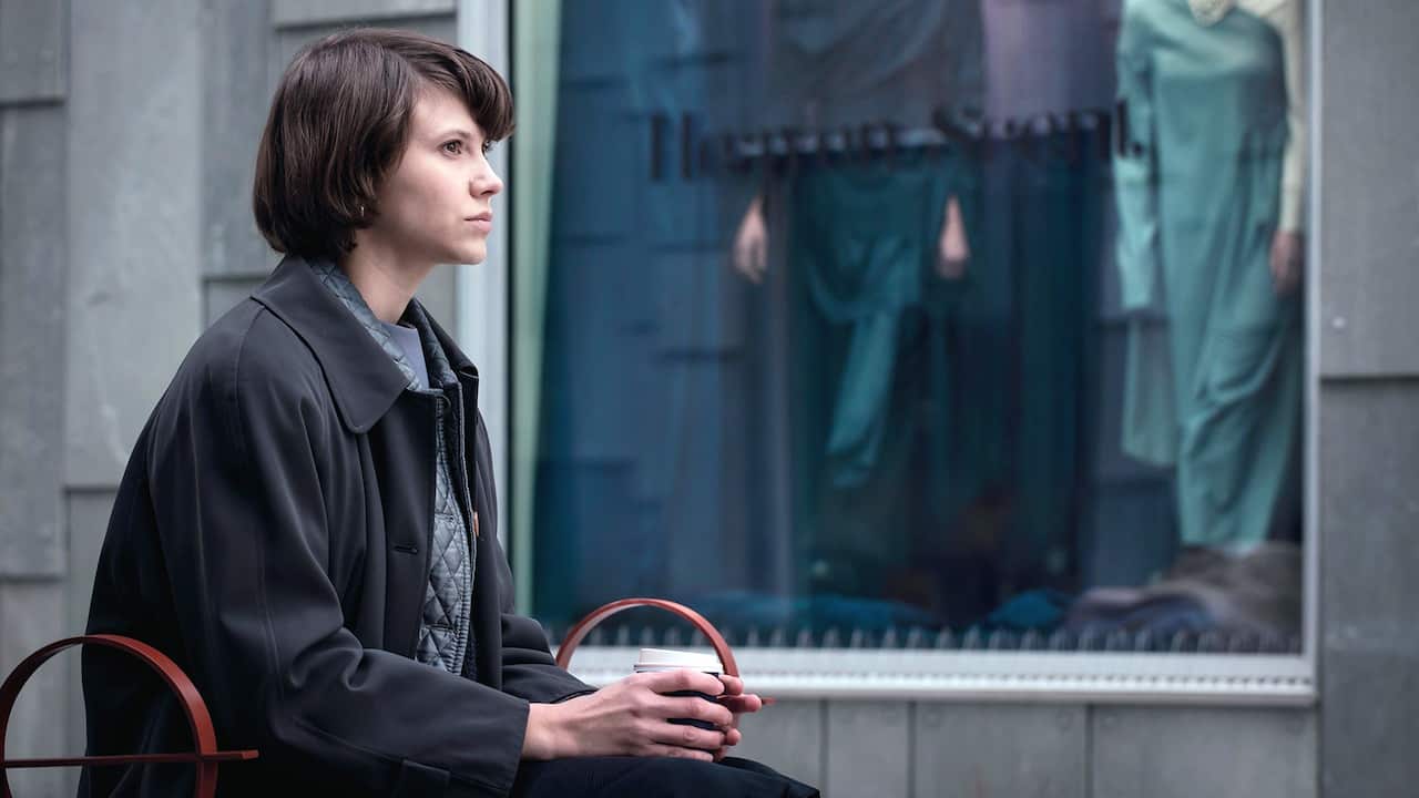 A woman in dark toned clothing sits, sideways, in front of a window in a grey building. She holds a takeaway coffee cup in her hands and stares pensively to into the distance. 