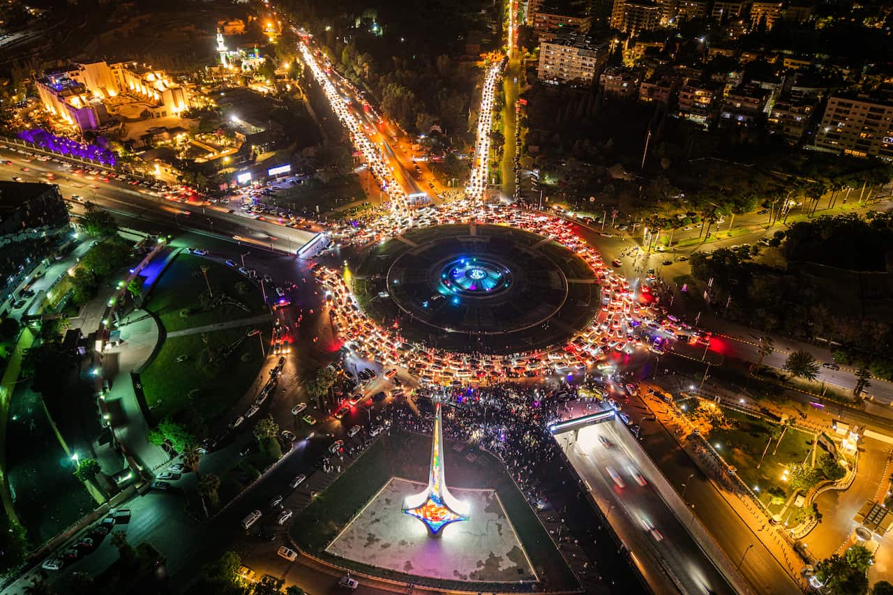 An aerial view of a busy, lit-up central square connecting to highways and roads leading to various areas in a city. There are buildings and vehicles visible at a distance.