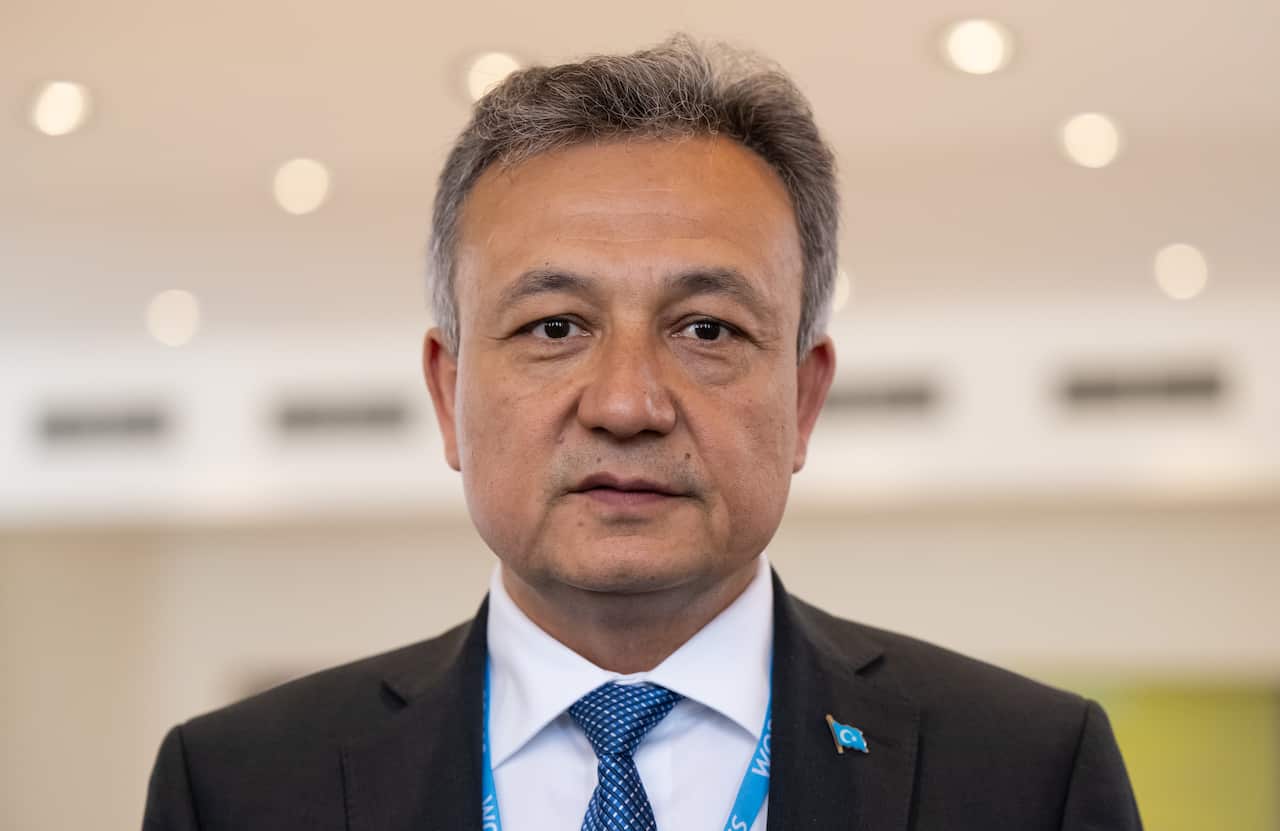 Dolkun Isa stands in a suit, with a Uyghur flag badge pinned to his collar.