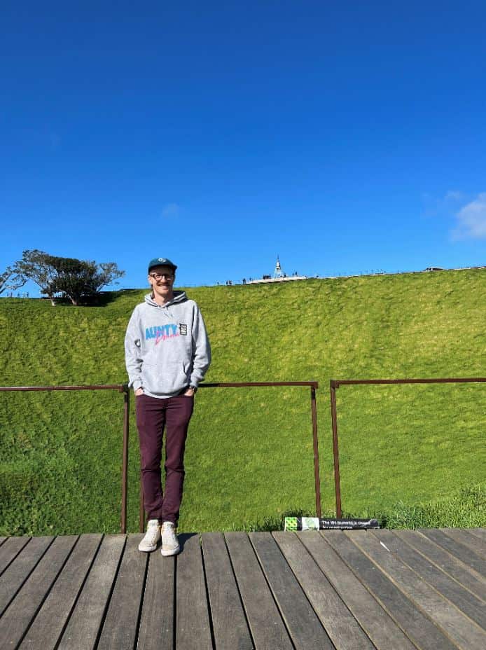 A young man leans on a wire fence in front of a lawn 