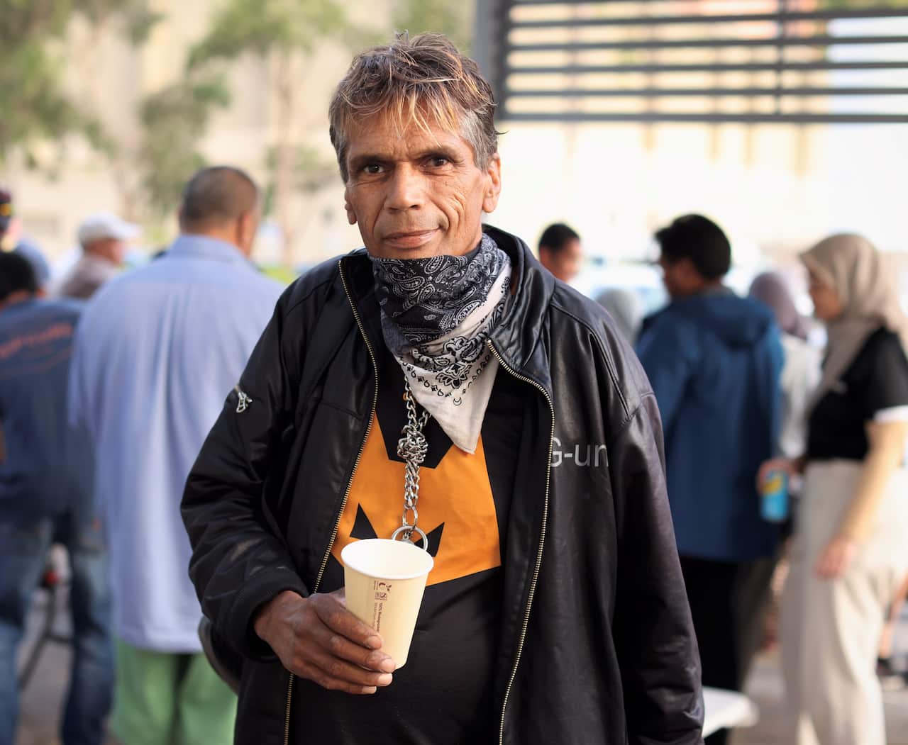 A man in a black jacket with a scarf around his neck stands holding a coffee cup.