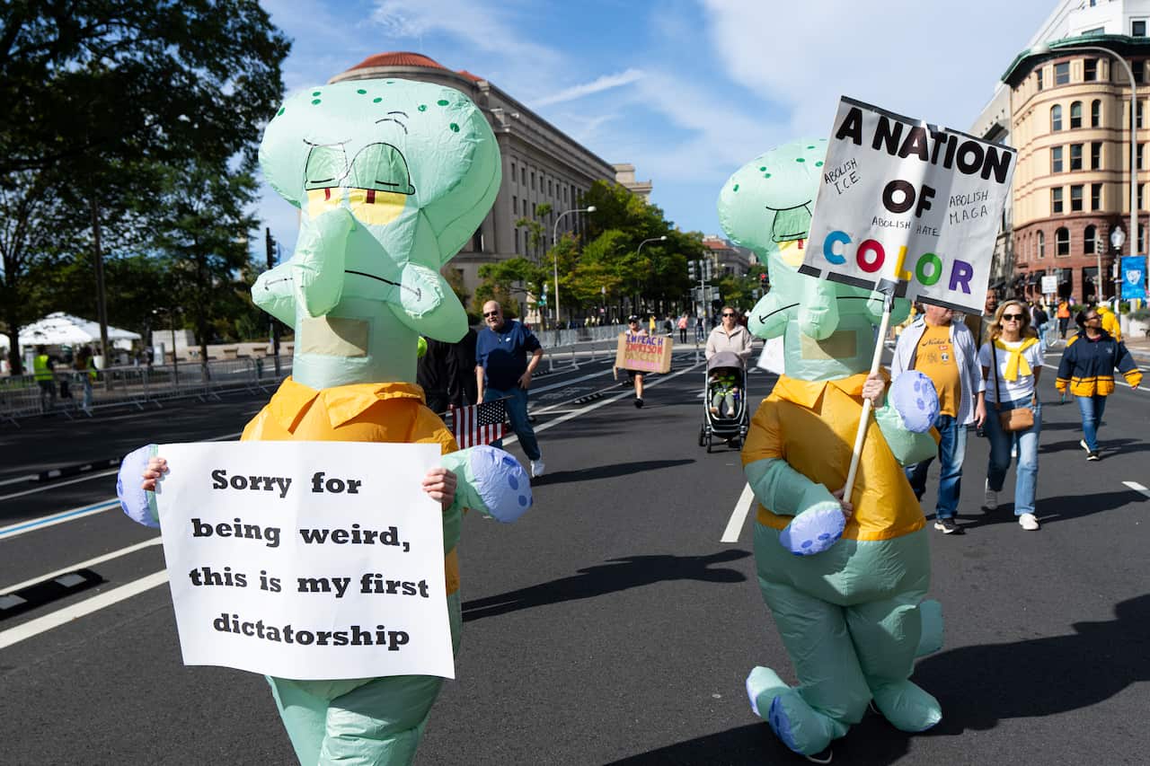 Two people in full-body, inflatable Squidward costumes from SpongeBob SquarePants walk down a city street during a protest. The person on the left holds a sign that reads, "Sorry for being weird, this is my first dictatorship," while the one on the right holds a sign with a stylized rainbow border that declares, "A NATION OF COLOR."