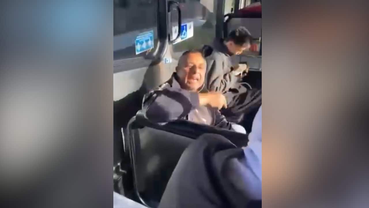 A man seated on a public bus speaks while other passengers sit nearby in footage recorded inside the vehicle.