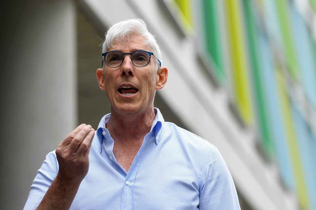 A man with grey hair and a white striped shirt speaks, gesturing with his hand.