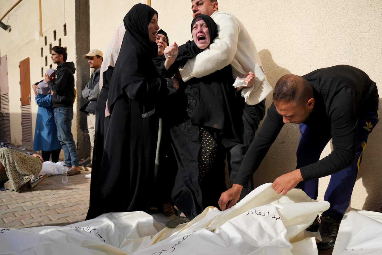 A woman crying while being held as white body bags are on the ground.
