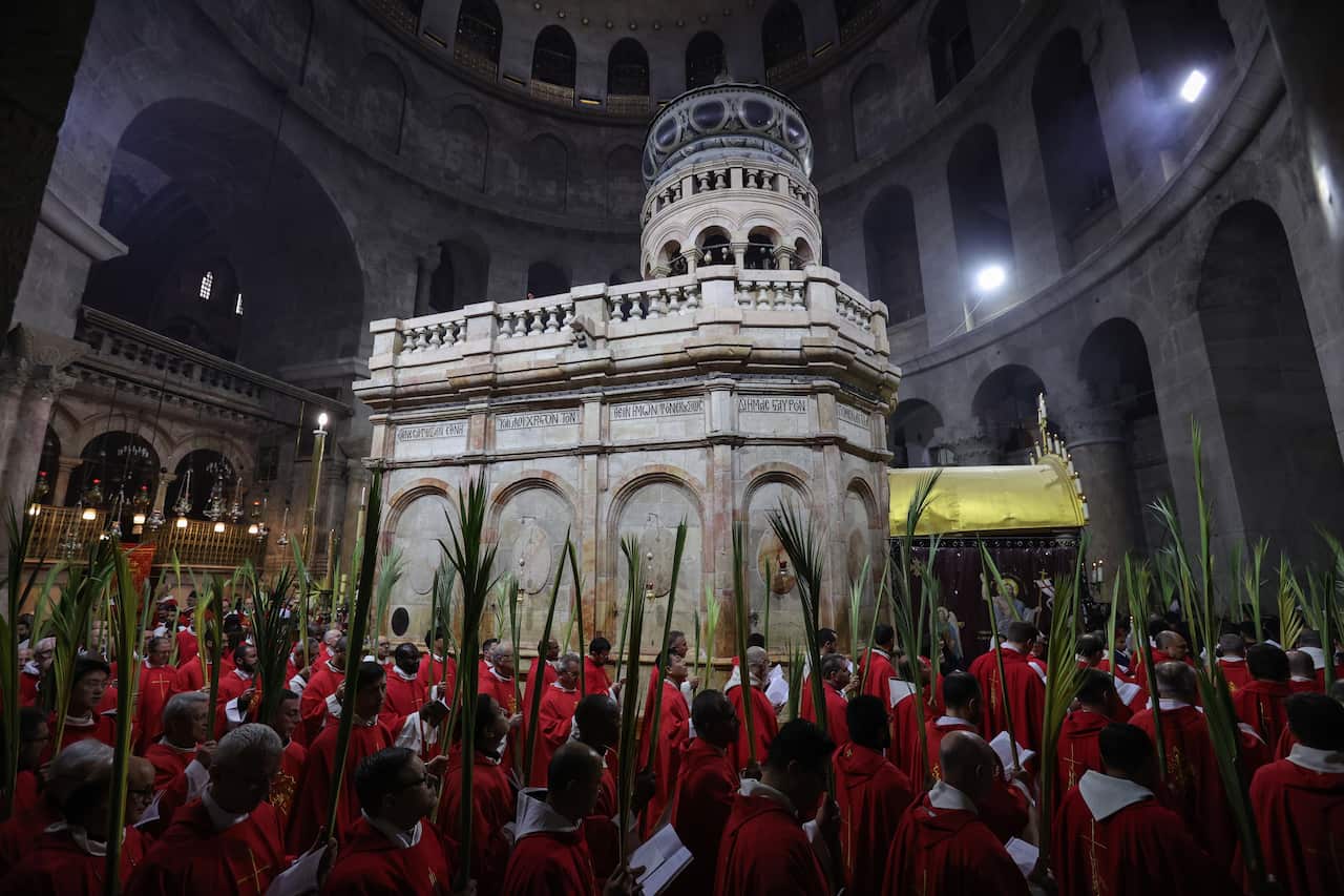 A large group of clergy in red robes carry palm fronds during a procession around a large stone shrine inside a large, ancient building.