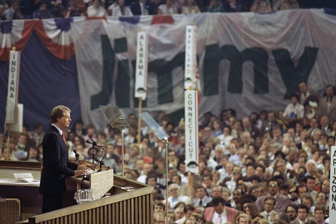 A man in a dark suit addresses a crowd as he stands at a podium on a raised platform.