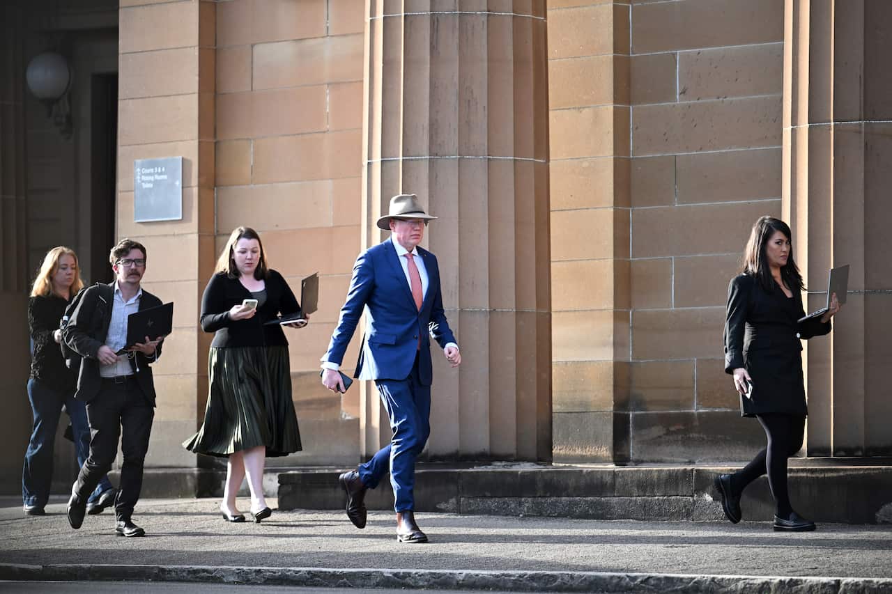 A man in a blue suit and a hat walks outside a courthouse. There is a woman in front of him and people walking behind him. 