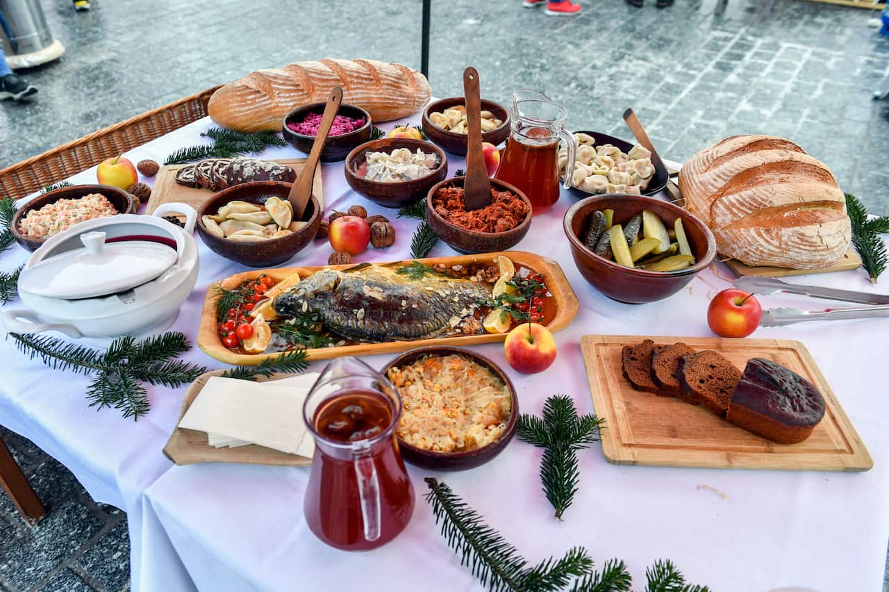 An outdoor table is laden with a variety of traditional Eastern European Christmas dishes, including a whole roasted fish, bowls of dumplings and salads, fresh loaves of bread, and pitchers of dark juice.