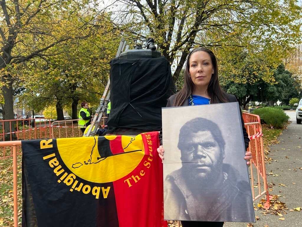 A woman holding a large photograph showing an Aboriginal man's face in front of a toppled statue.