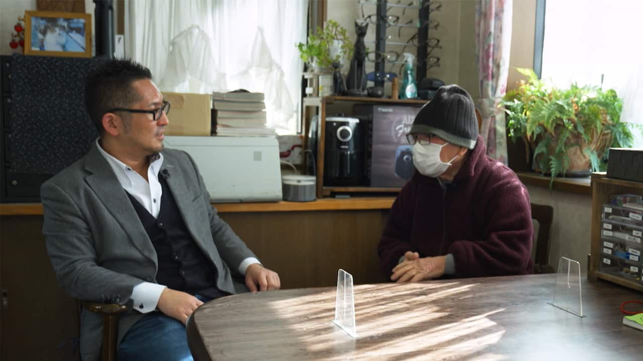 Two men sit next to each other talking in an office. On the left is a middle-aged Japanese man wearing a grey suit and white shirt. Opposite him is an elderly man wearing a grey beanie and a white medical mask
