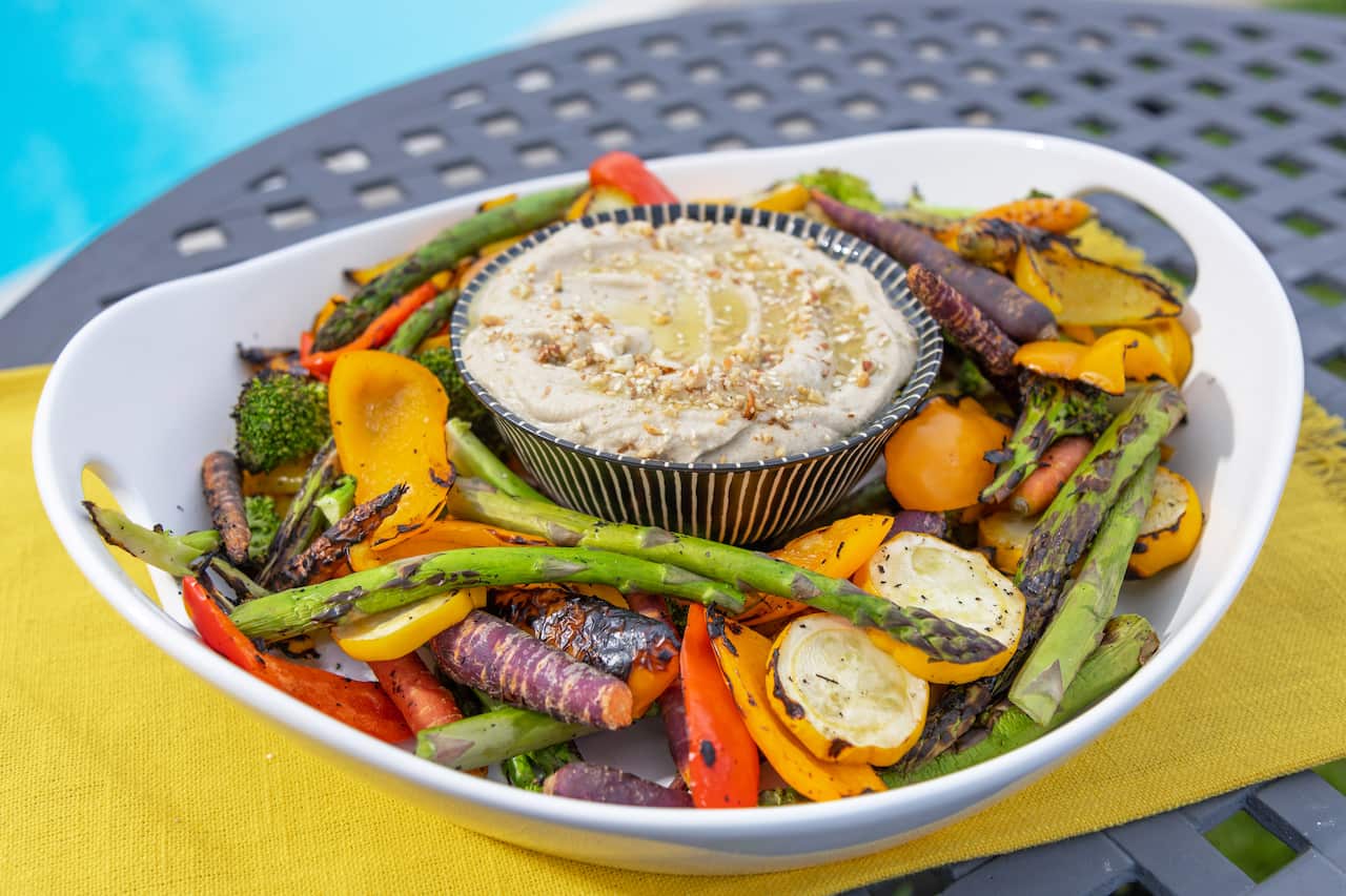 In a wide white bowl holds a colourful array of grilled vegetables and a small striped bowl filled with creamy dip.