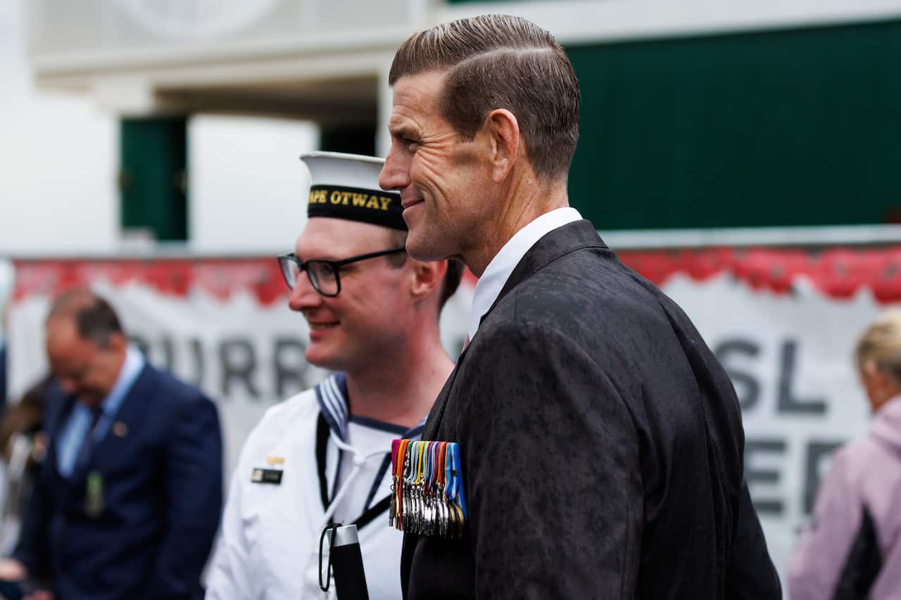 A tall man in a suit in the foreground standing next to man with a navy uniform in the background.